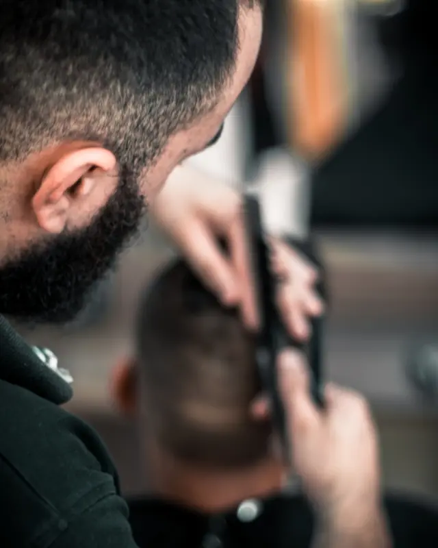 Un barbero con barba repasa con la máquina la nuca de un cliente.